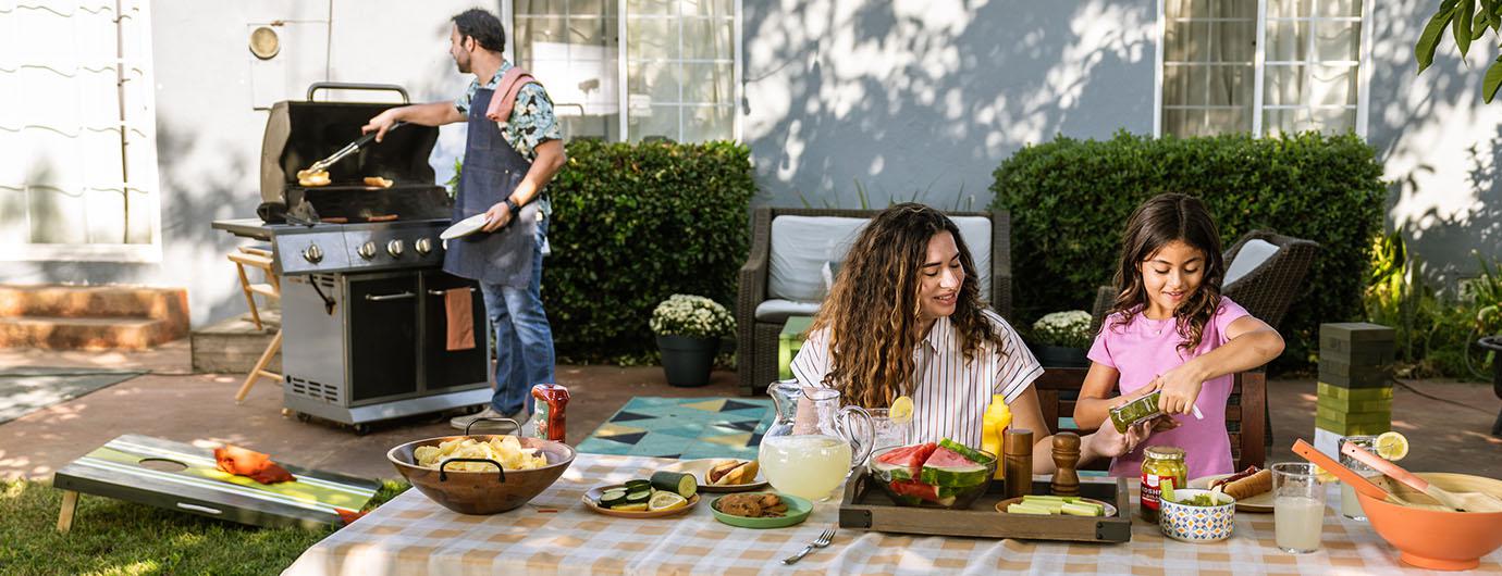 man grilling, women and child sitting at table full of food