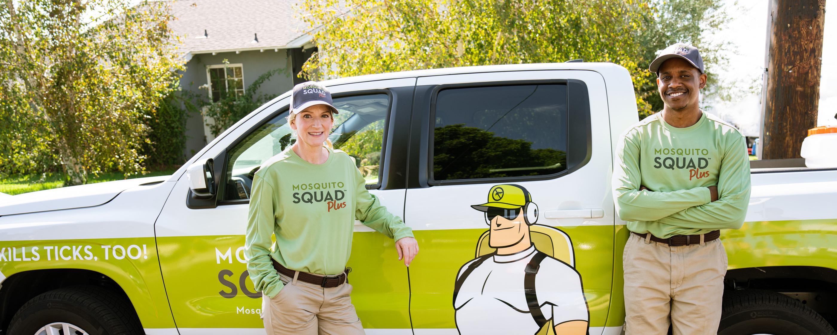 Two Mosquito Squad technicians  in green uniforms and hats, equipped with sprayers, in front of a brick house.